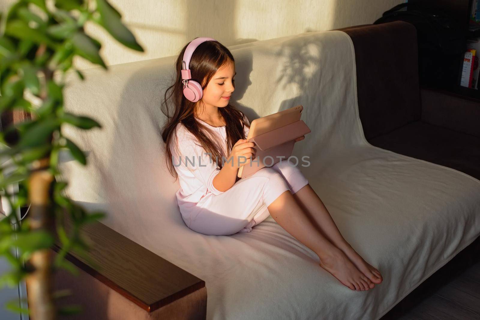 A little beautiful girl in pink home clothes and pink headphones sits on the couch, looks into a digital tablet.