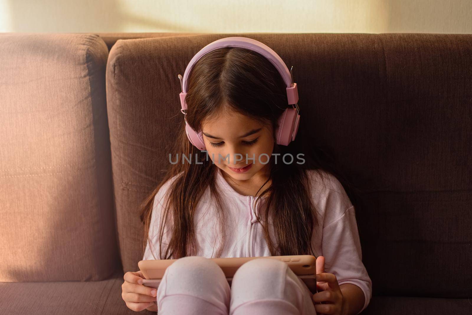 A little smiling girl in pink headphones sits on the brown couch, looks into a digital tablet.