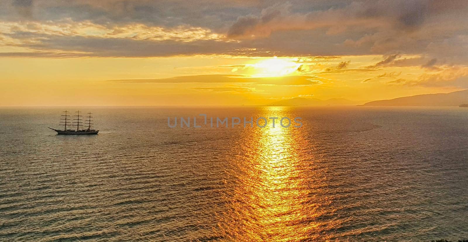 Romantic golden sunset on the sea with a yacht against the background of mountains and orange clouds, a calm scene of a warm peaceful evening.