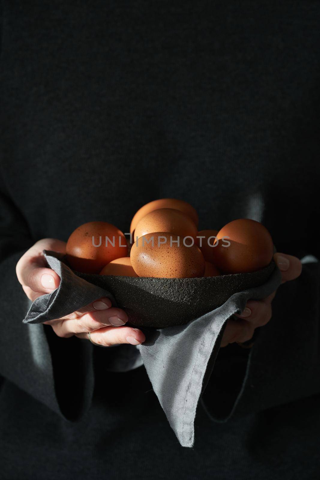 Unusual Easter on a dark background. A bowl of brown eggs with hands. Darkness, rays of sunlight shine on the eggs. The concept of a new life, rebirth. Rustic style. Vertical, copy space