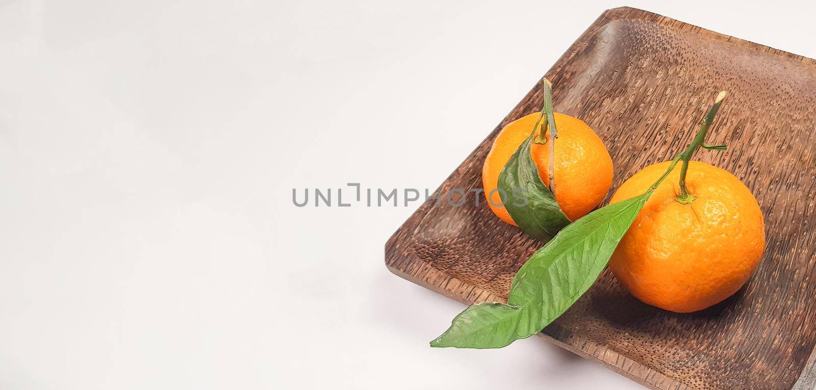 Two fresh ripe tangerines with leaves and branches on a dark square wooden plate, close-up, space for text on the left.
