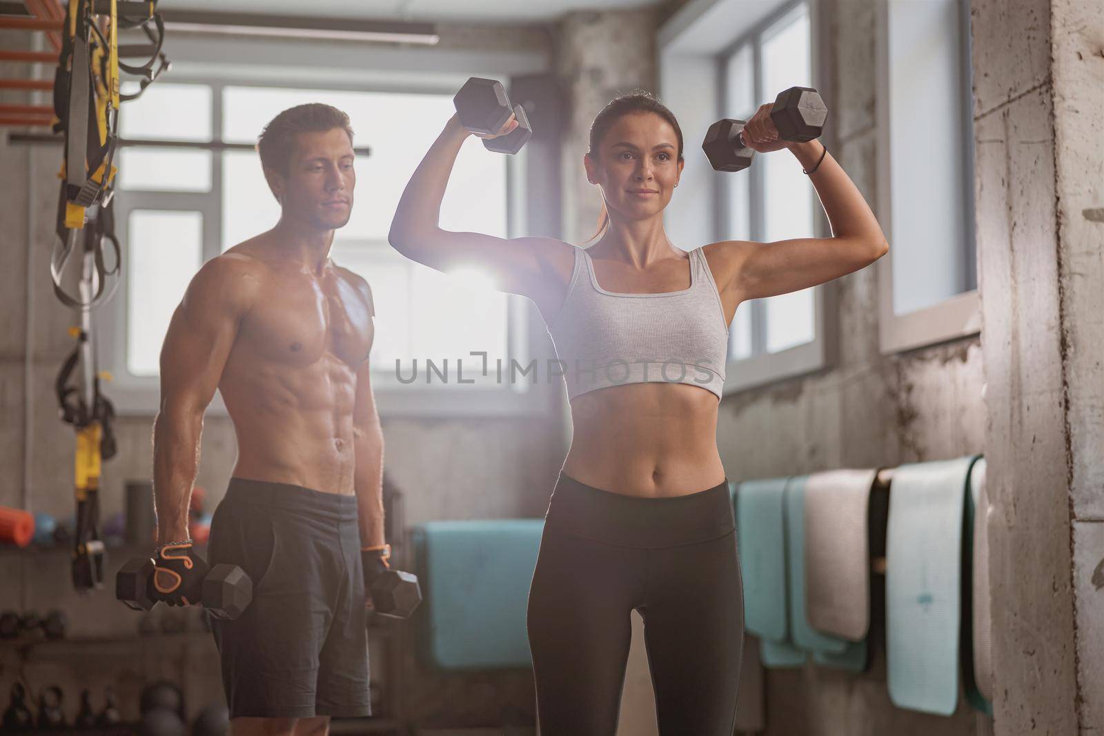 Slender pretty woman holding dumbbells and raising them, performing a sports exercise with strong man in the gym