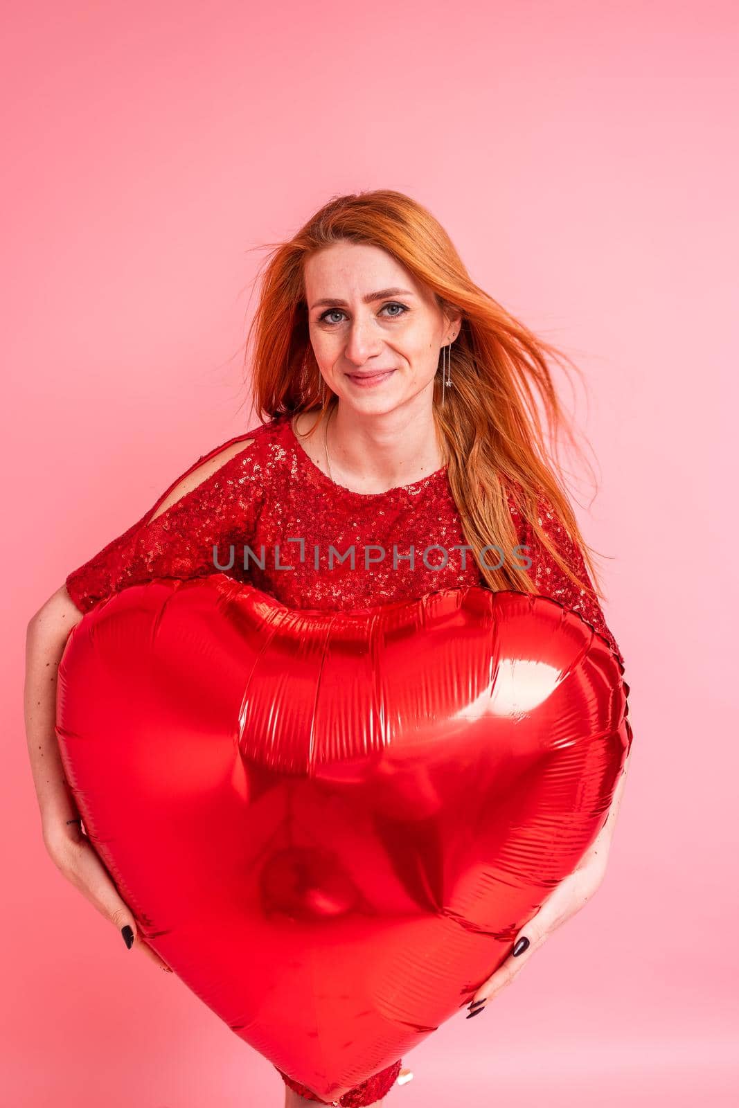 Beautiful redhead girl with red heart baloon posing. Happy Valentine's Day concept. Studio photo of beautiful ginger girl dancing on pink background.