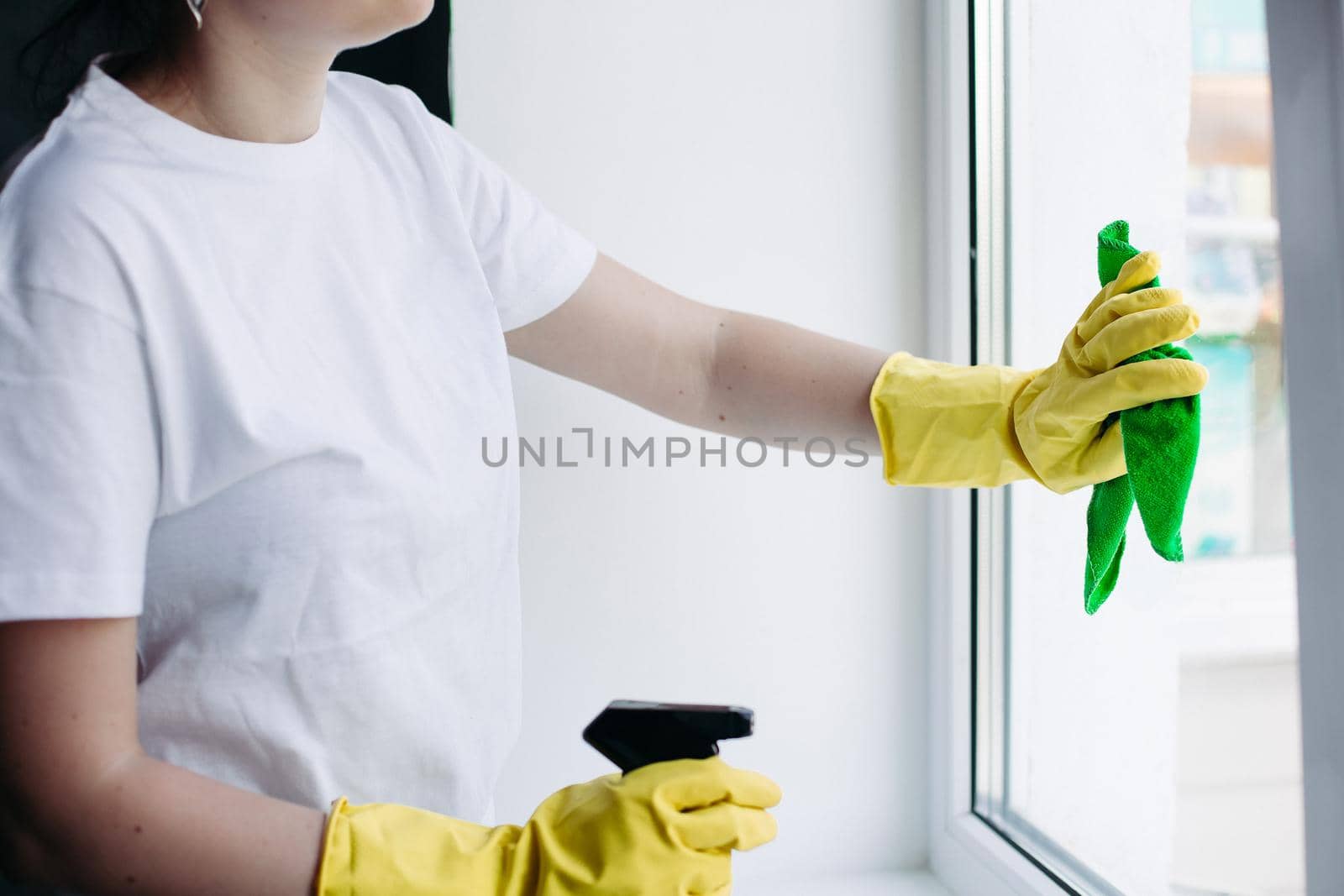 Crop of incognito brunette housewife cleaning dirty window. Housemaid wearing white t shirt and yellow protective gloves wiping glass with rag. Concept of housework and apartment service.