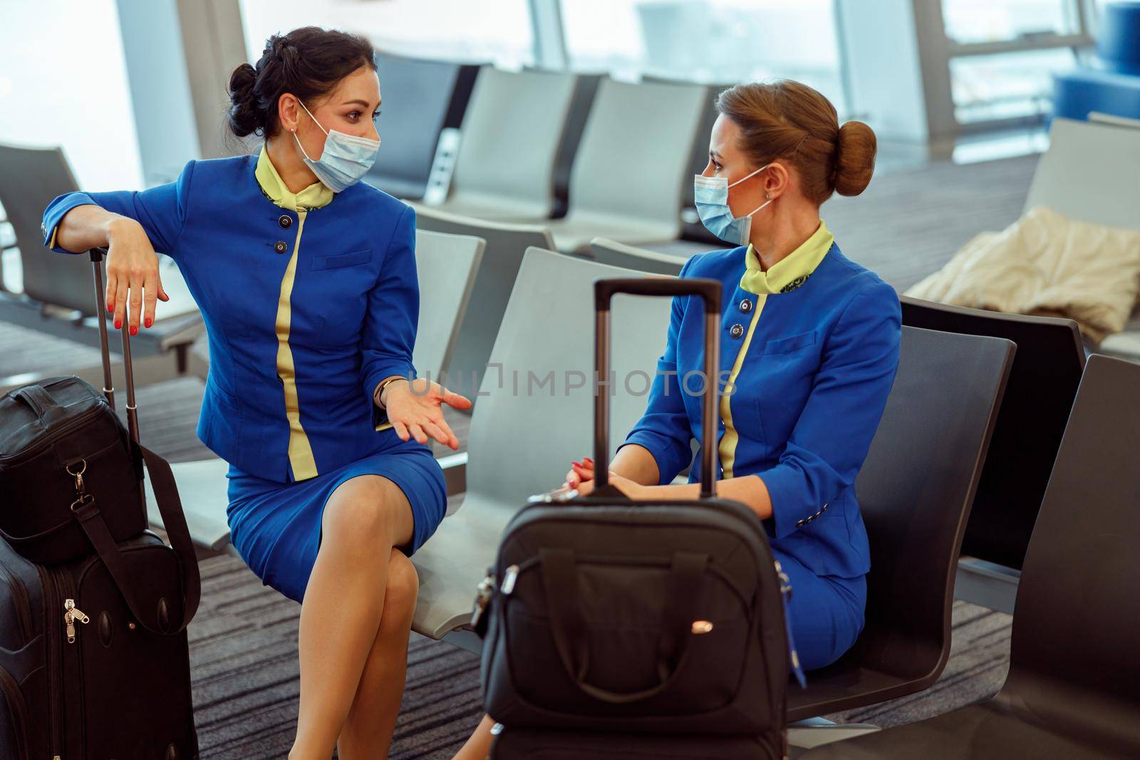 Flight attendants in medical masks sitting on chairs at airport by Yaroslav_astakhov