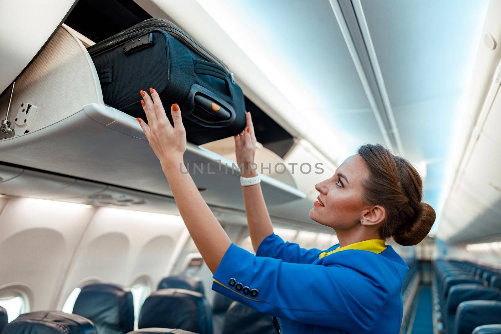 Stewardess putting travel suitcase in overhead luggage bin in aircraft by Yaroslav_astakhov