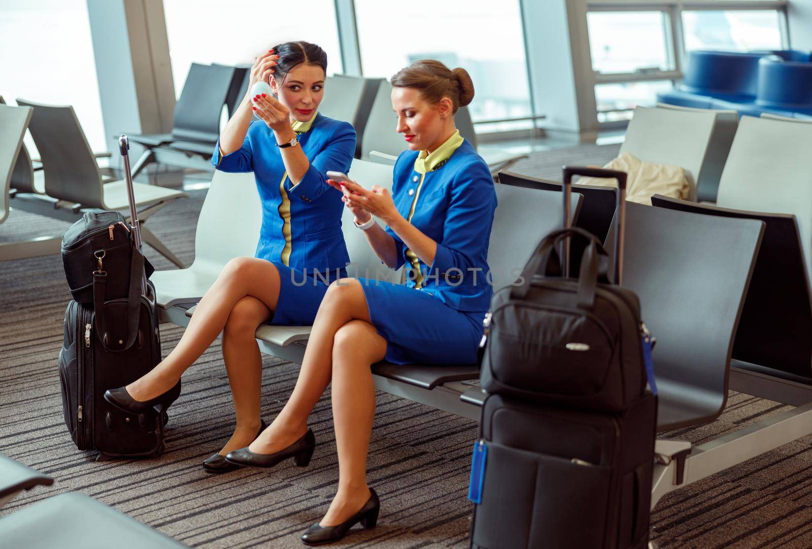 Female flight attendant sitting on chair and looking in travel mirror while colleague using smartphone in departure lounge