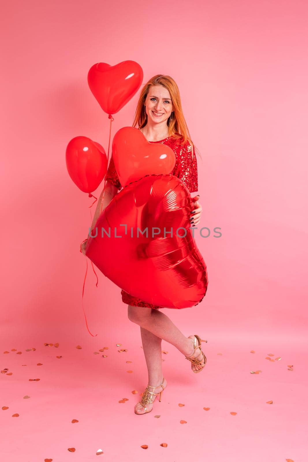 Beautiful redhead girl with red heart baloon posing. Happy Valentine's Day concept. Studio photo of beautiful ginger girl dancing on pink background.