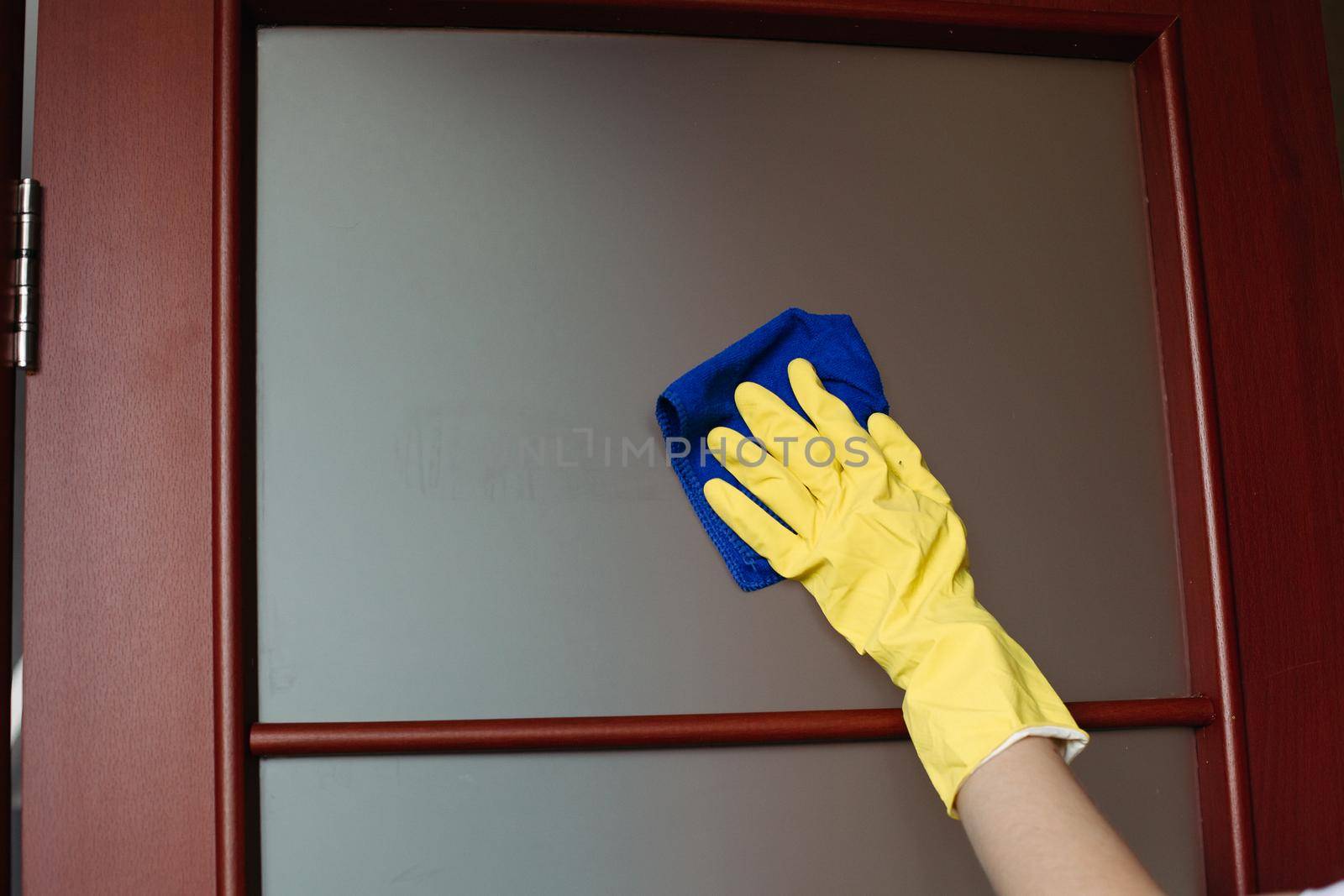 Crop of housewife hands wearing yellow protective gloves, cleaning dirty window in doors. Housemaid wiping glass with rag. Concept of housework and service.