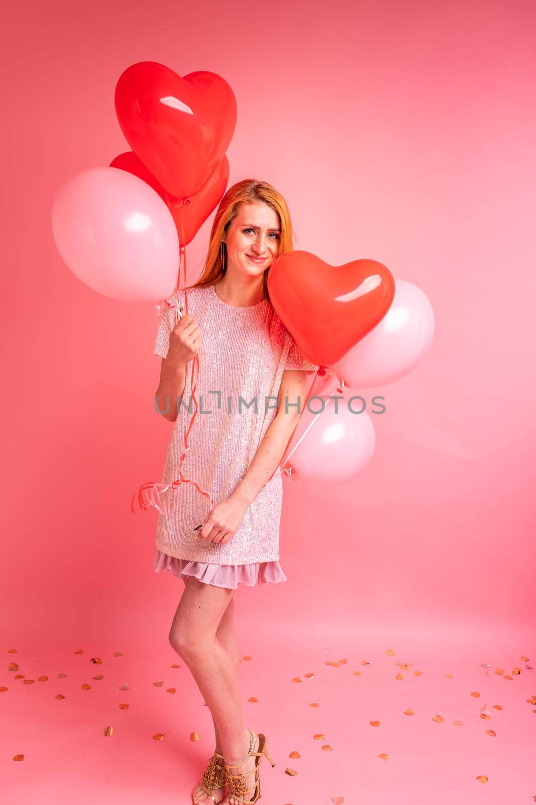 Beautiful redhead girl with red heart baloon posing. Happy Valentine's Day concept. Studio photo of beautiful ginger girl dancing on pink background.