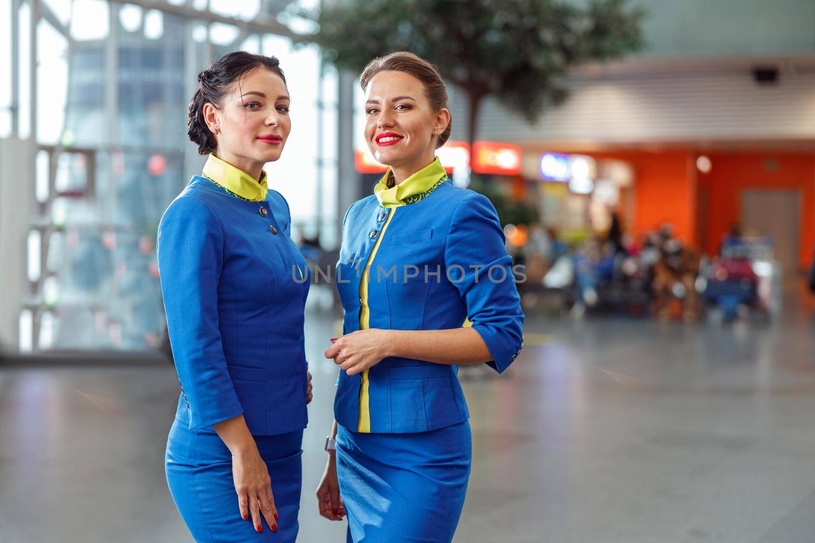 Joyful women stewardesses in aviation uniform looking at camera and smiling while standing in passenger flight terminal