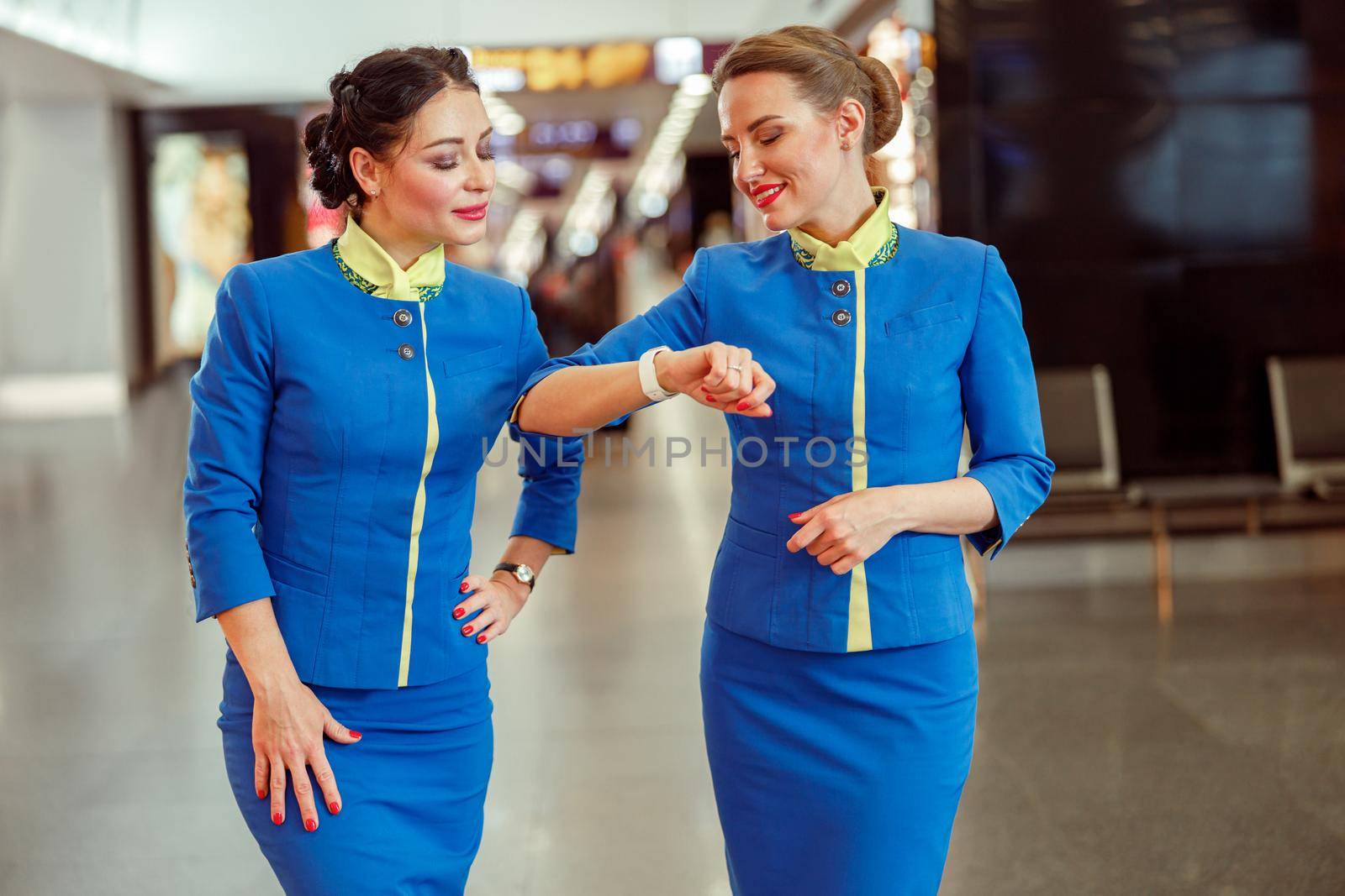 Joyful woman stewardess in air hostess uniform looking at watch and smiling while waiting for plane with colleague in airport terminal