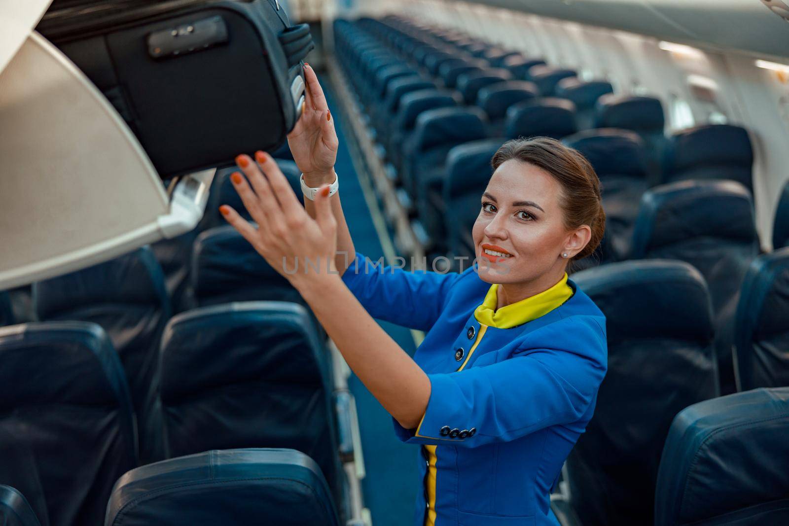 Smiling woman flight attendant placing travel suitcase in overhead baggage compartment while standing in aircraft passenger salon