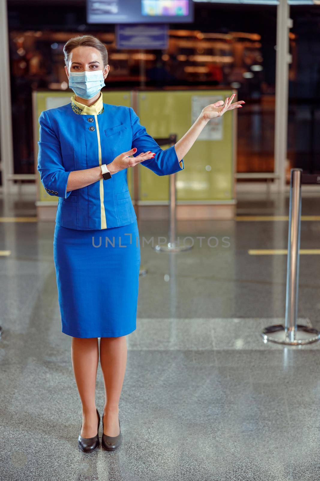 Female flight attendant wearing protective face mask and air hostess uniform while pointing at something in airport terminal