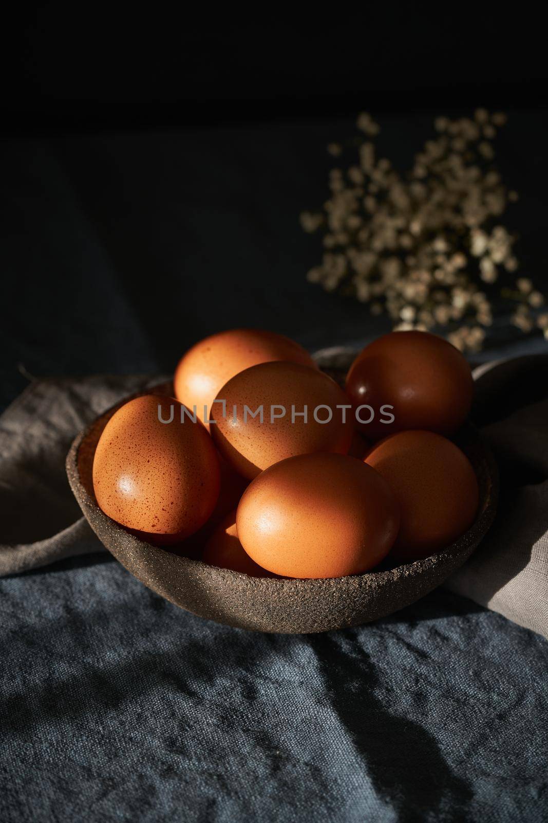 Unusual Easter on dark background. Bowl of brown eggs on dark blue table, flowers. Darkness, rays of sunlight shine on eggs. Concept of new life, rebirth. Side view, copy space, vertical