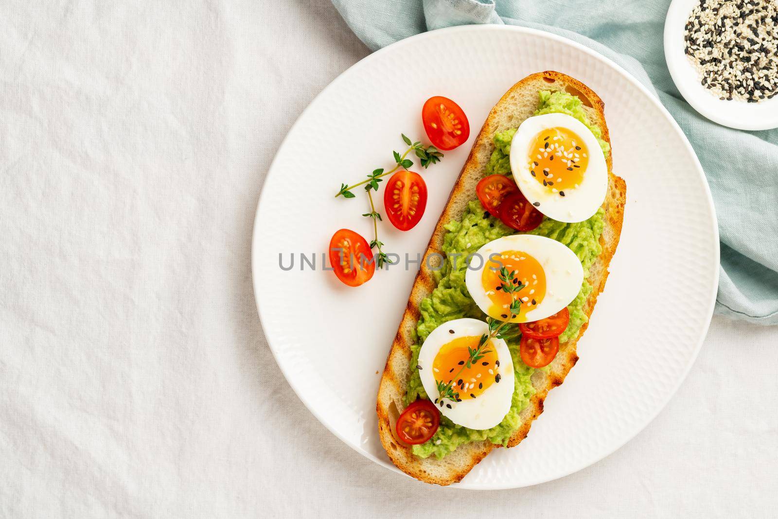 Avocado toast with toasted bread soft-boiled eggs with yellow yolk and tomatoes with herbs on white plate on light tablecloth