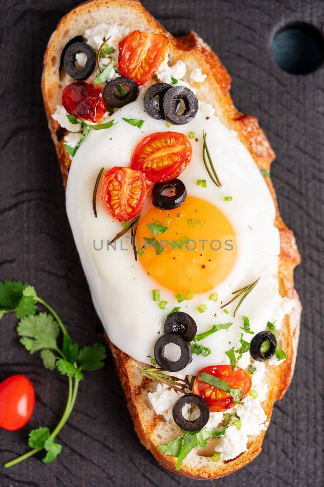 Toasted bread toast with fried eggs with yellow yolk and tomatoes, olives, sprinkled with herbs on dark wooden serving board on blue denim napkin, vertical, top view, close up