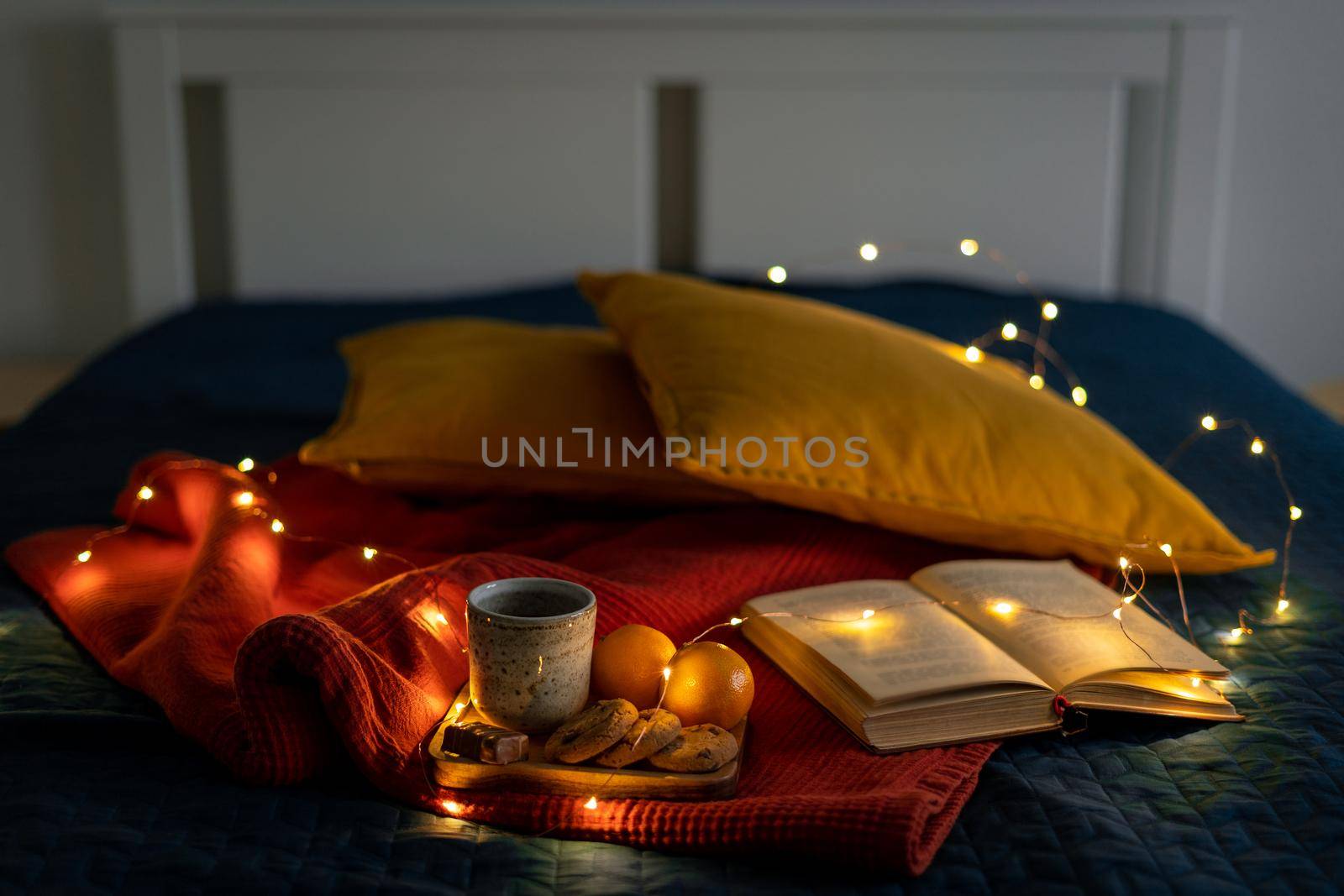 Cozy background for reading book relaxing and reflection. Stay home concept. Christmas decoration with garland lights, pillows and hot drink with cookies on bed. Dark evening twilight in bedroom