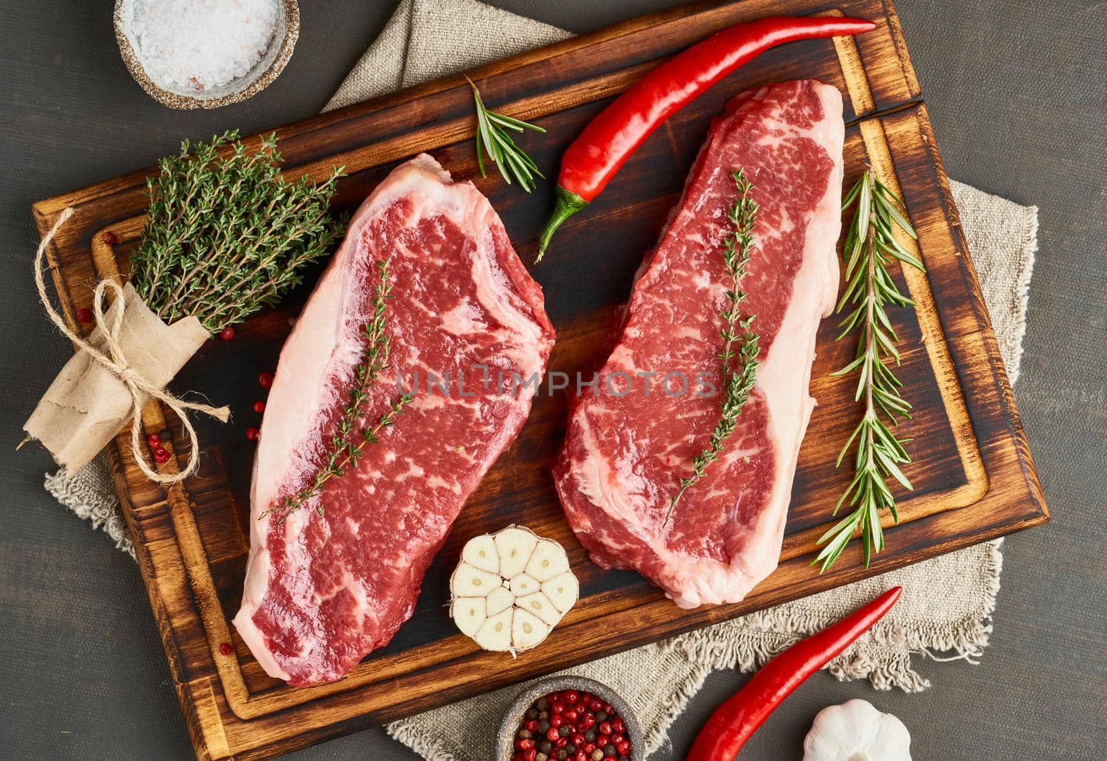 Big whole piece of raw beef meat, striploin on wooden cutting board on dark brown background. Seasoning steak with salt, thyme, garlic. Top view