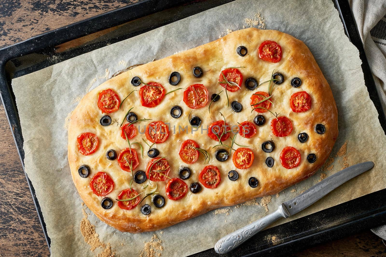 Focaccia, pizza, italian flat bread with tomatoes, olives and rosemary on tray on dark wooden rustic table.