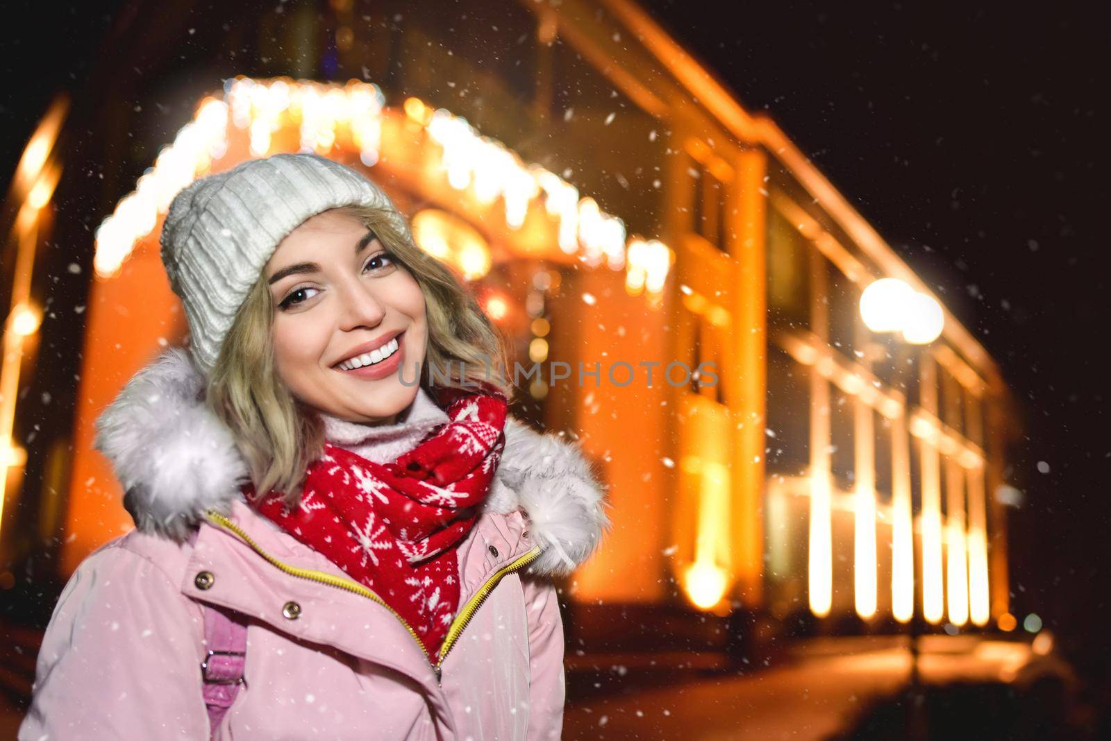 portrait of a charming woman on the snowy street, in a winter jacket, smiles happily around the evening lights of the city, she looks into the camera and shines with happiness