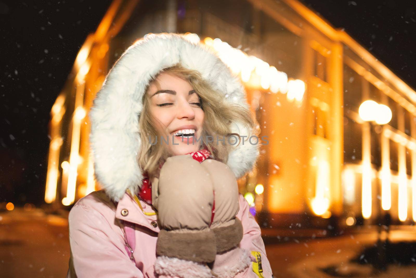 portrait of a charming woman on a snow-covered street, wearing a winter jacket and hoodie, smiling happily at the evening lights of the city, she glows with happiness with her eyes closed