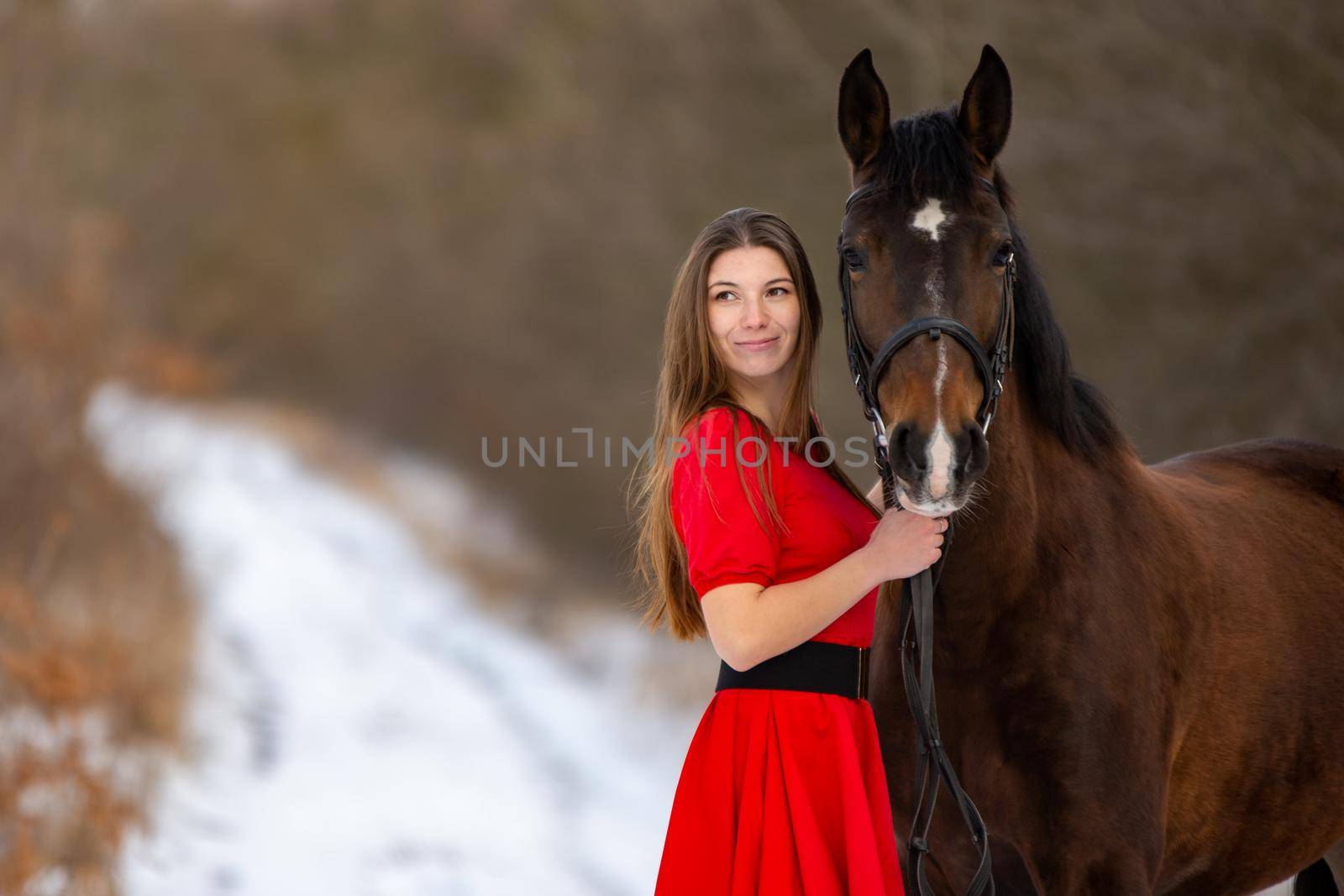 Portrait of a beautiful girl in a red dress standing with a horse on the background of a winter road a
