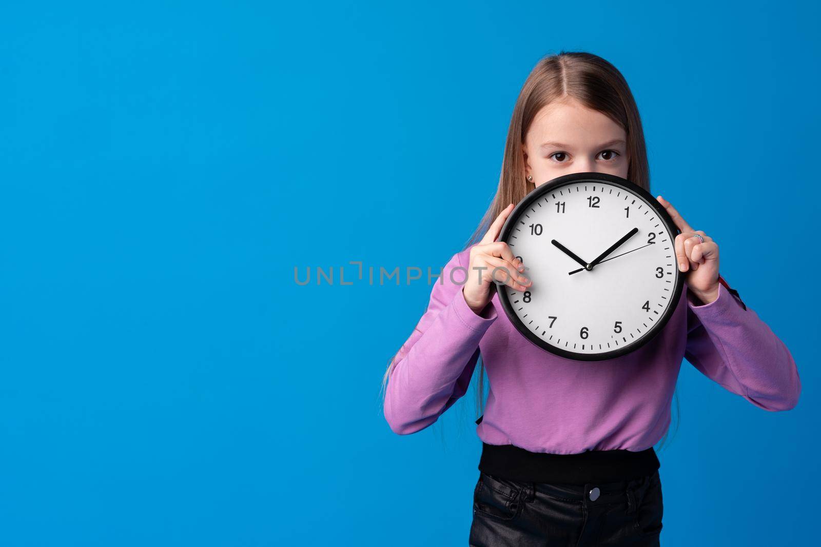 Beautiful teen girl holding wall clock over blue background, close up