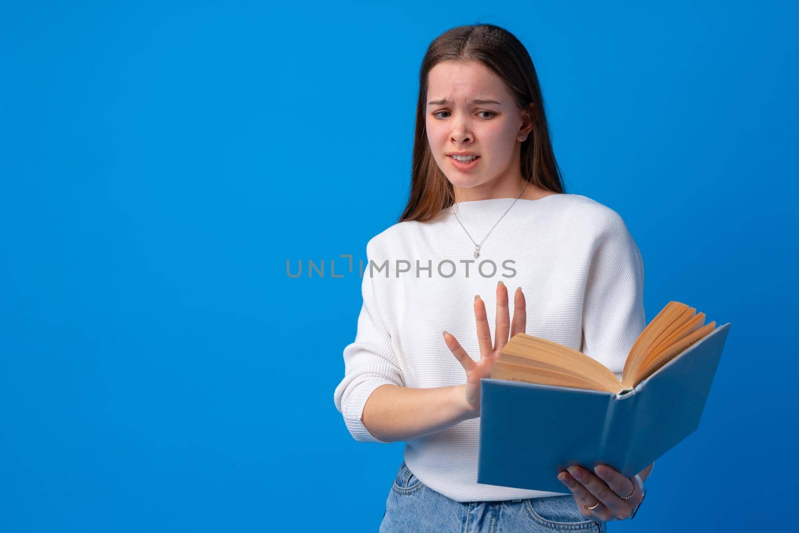Young beautiful woman holding book scared and amazed on blue background in studio, close up