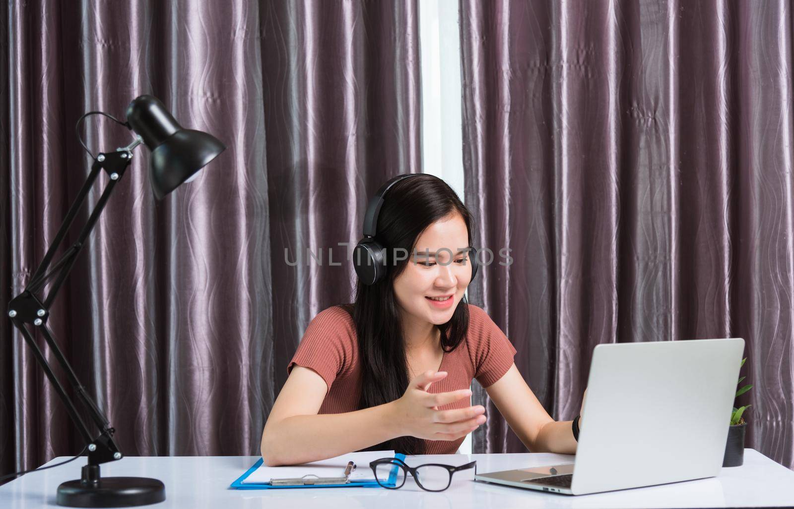 Work from home, Asian young businesswoman smile wearing headphones video conference call or facetime by laptop computer her raise his hand to explain to teammates on desk at home office