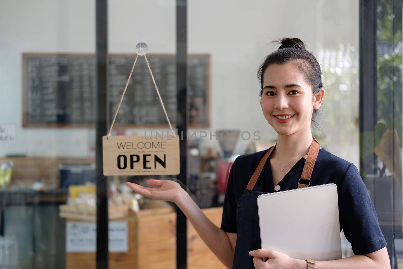 Beautiful asian young barista woman in apron holding tablet and standing in front of the door of cafe with open sign board. Business owner startup SME entrepreneur concept