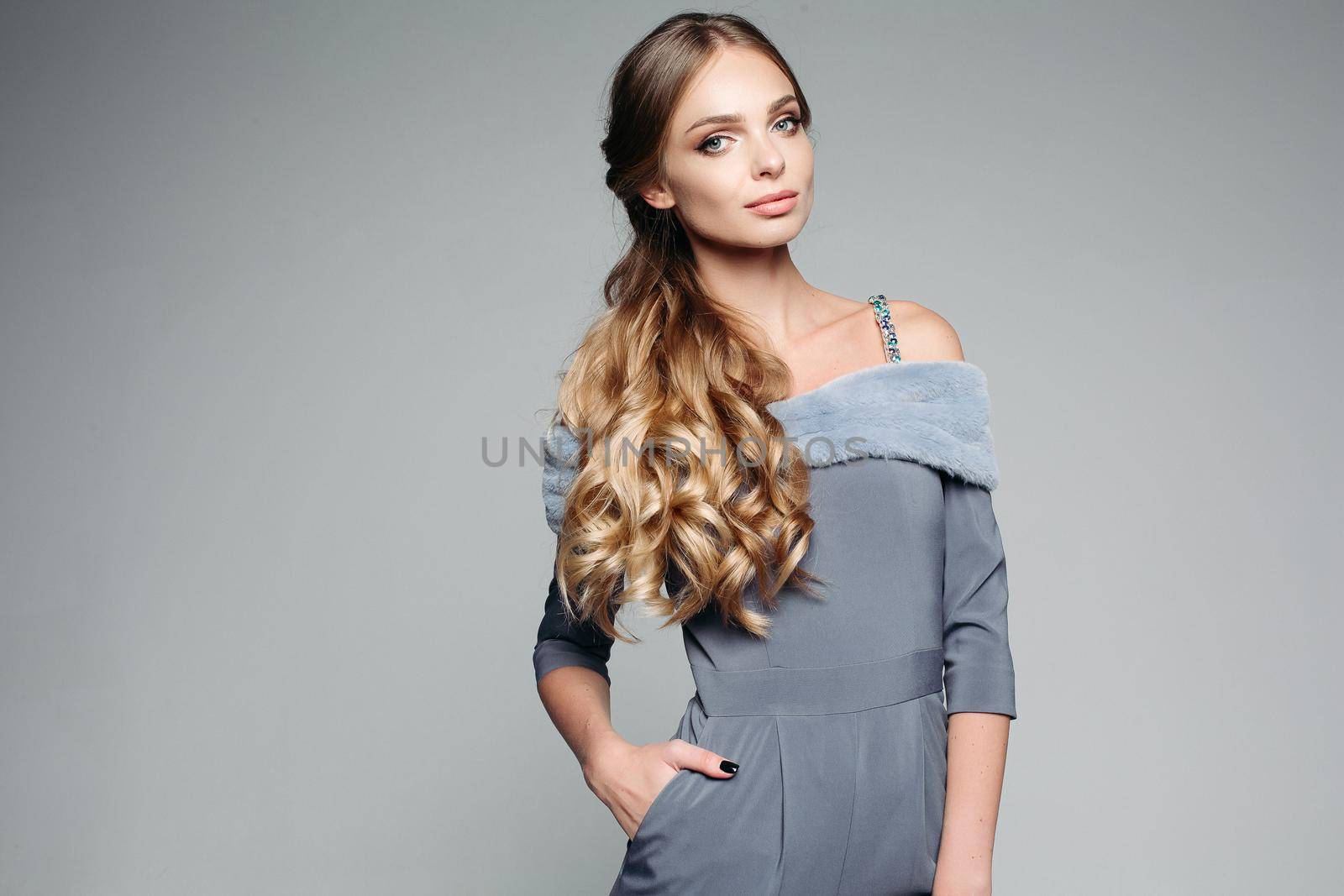 Gorgeous blondie woman wearing in gray outfit holding hand in pocket, seriously looking at camera. Seductive girl with perfect skin, curly hair, professional make up, after beauty salon.