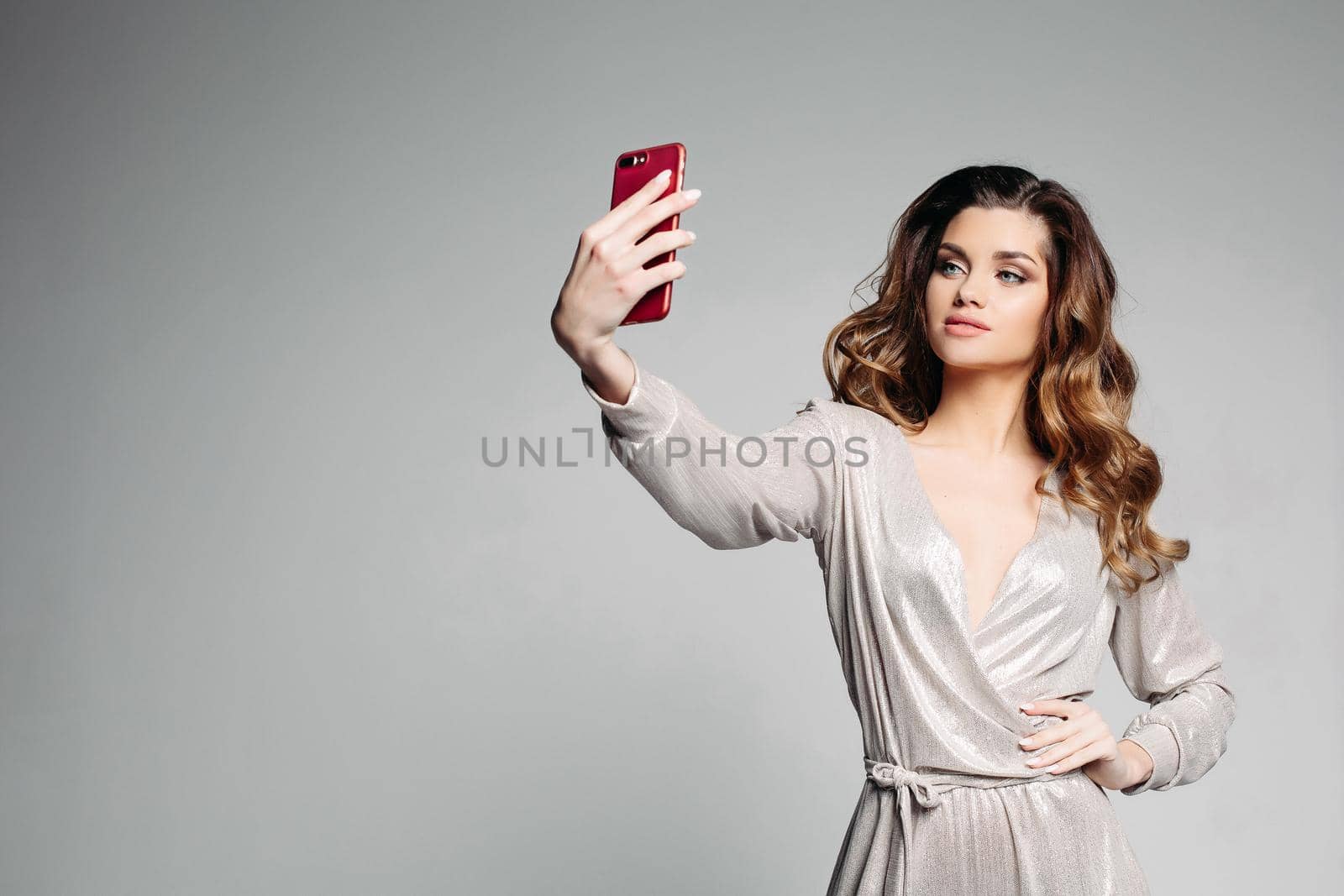 Stylish and attractive woman confident posing with hand on hips and taking self portrait at call phone. Sudactive female model in silver dress taking photo at smartphone. Concept of style.