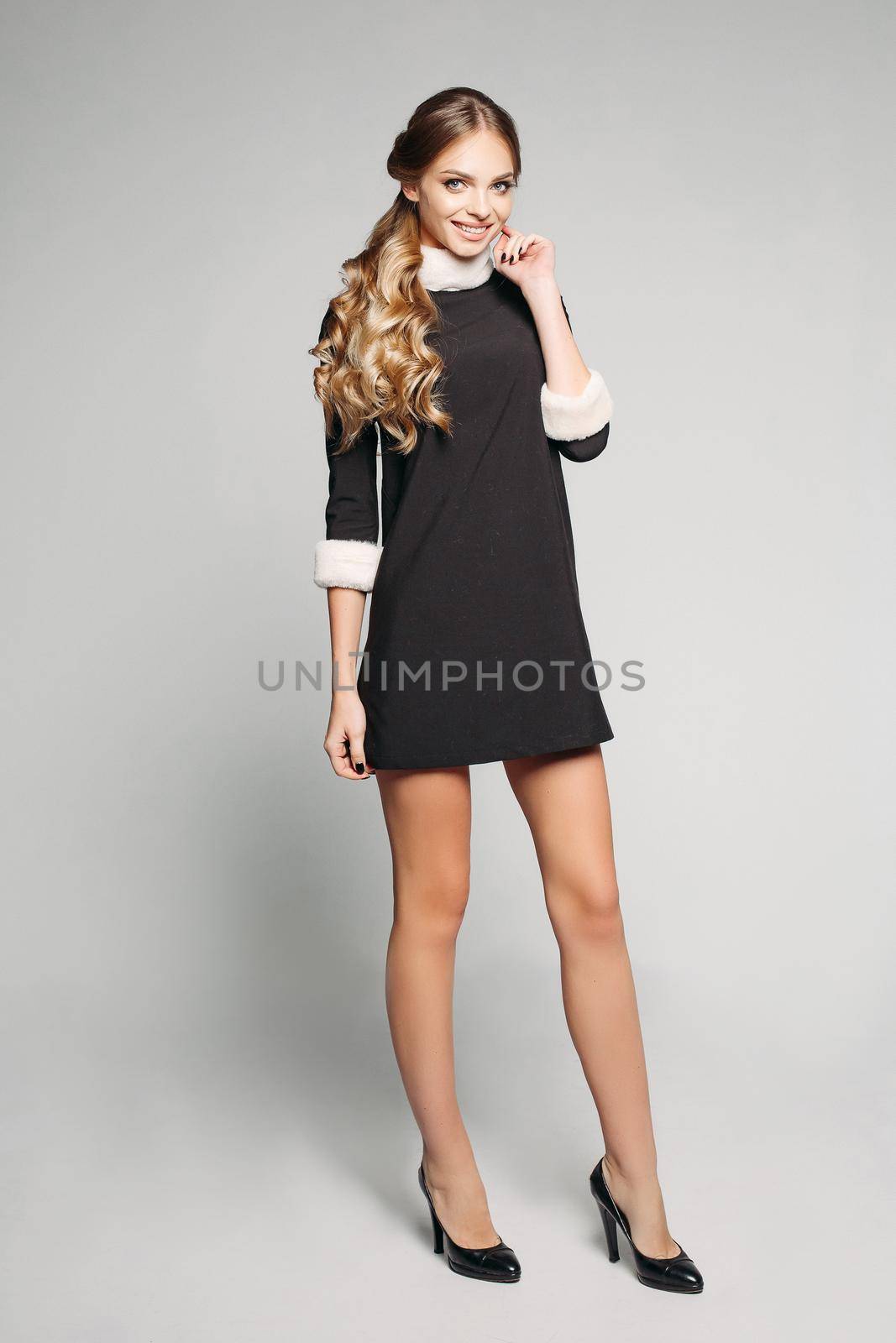 Full length of gorgeous smiling model with long wavy hair in tail wearing little black dress with fur trimming and high heels. Isolate on white background.