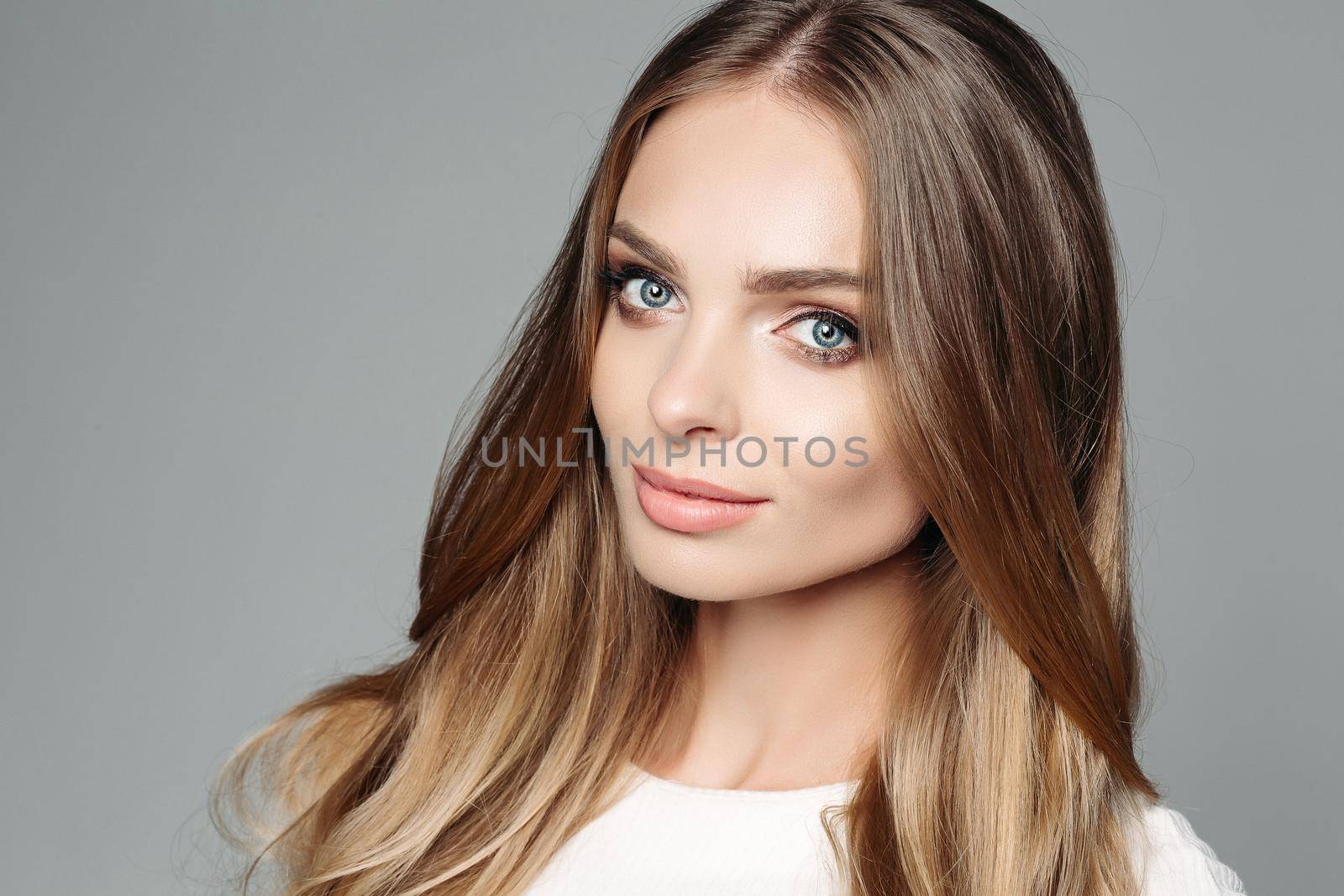 Studio portrait of an attractive blond woman with long thick hair and low-key makeup, wearing an elegant white blouse, cute looks at the camera on a gray background. Isolate. The concept of beauty.