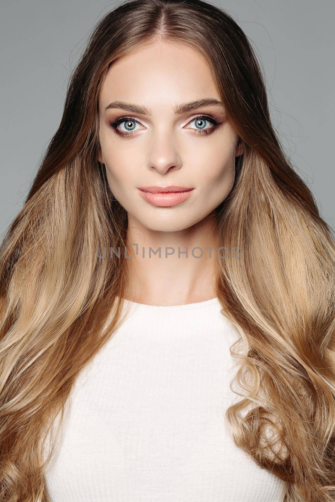 Studio portrait of an attractive blond woman with long thick hair and low-key makeup, wearing an elegant white blouse, cute looks at the camera on a gray background. Isolate. The concept of beauty.