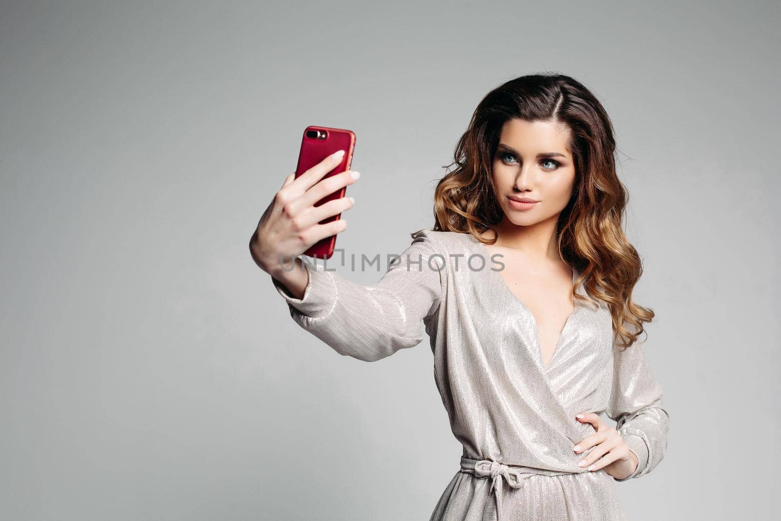 Stylish and attractive woman confident posing with hand on hips and taking self portrait at call phone. Sudactive female model in silver dress taking photo at smartphone. Concept of style.