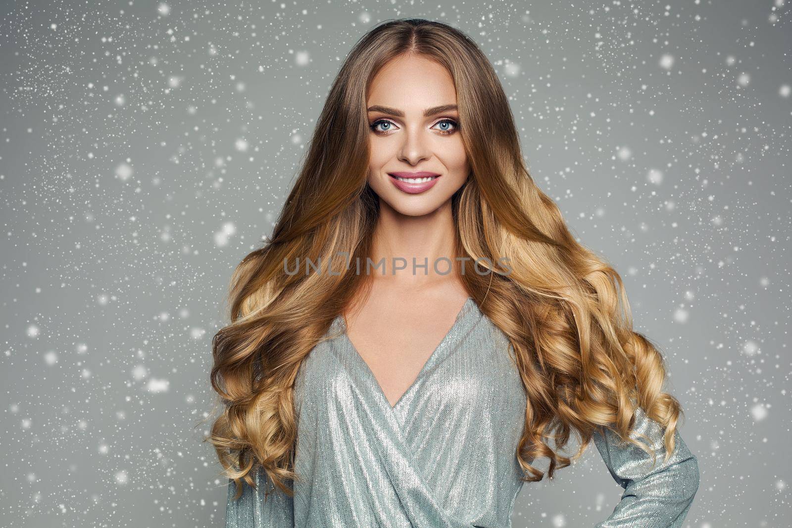 Studio portrait of attractive blonde woman with long thick hair and professional make-up wearing elegant silver blouse smiling at camera over grey background. Isolate. Beauty concept.