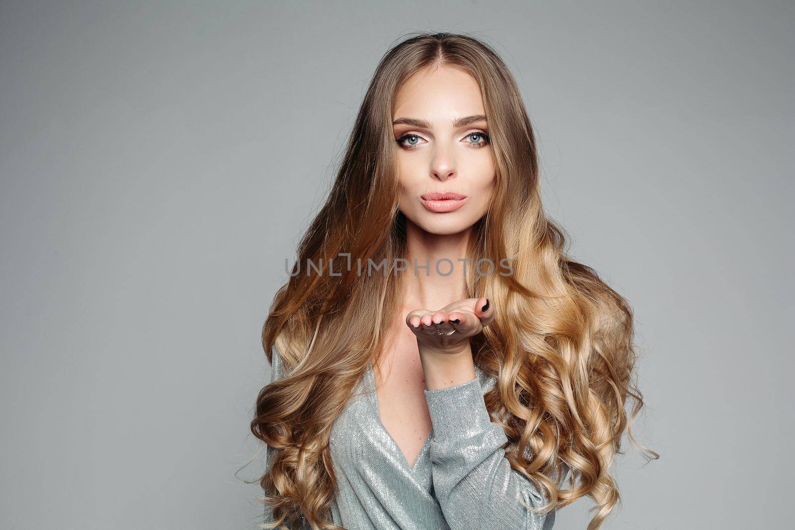 Studio portrait of an attractive blond woman with long thick hair and professional make-up wearing an elegant silver blouse, an air kiss on the camera on a gray background. Isolate. The concept of beauty.