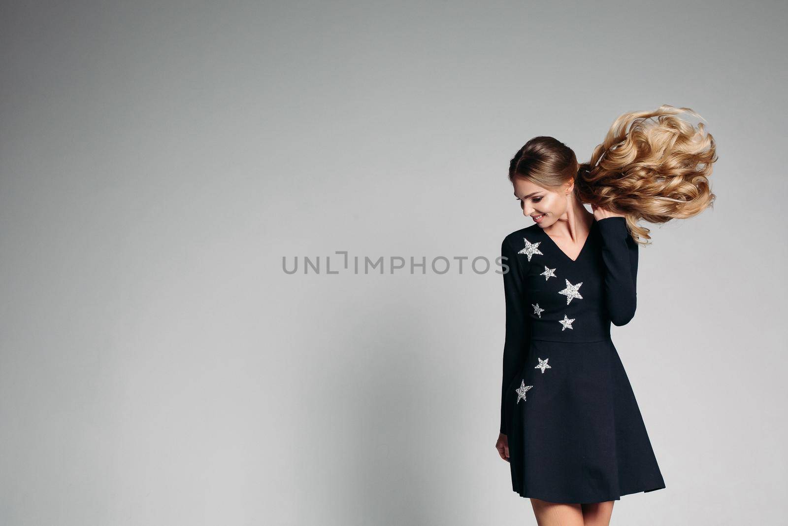 Portrait of beautiful positivity woman with long curly hair and shining eyes, wearing in fashionable black dress with stars, jumping in grey studio. Stylish woman dancing and smiling.