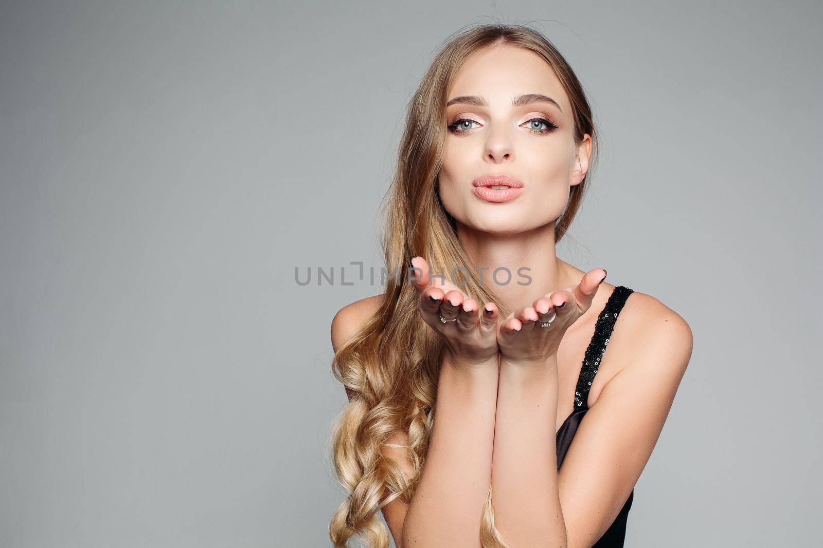 Studio portrait of an attractive blond woman with long thick hair and professional make-up wearing an elegant silver blouse, an air kiss on the camera on a gray background. Isolate. The concept of beauty.