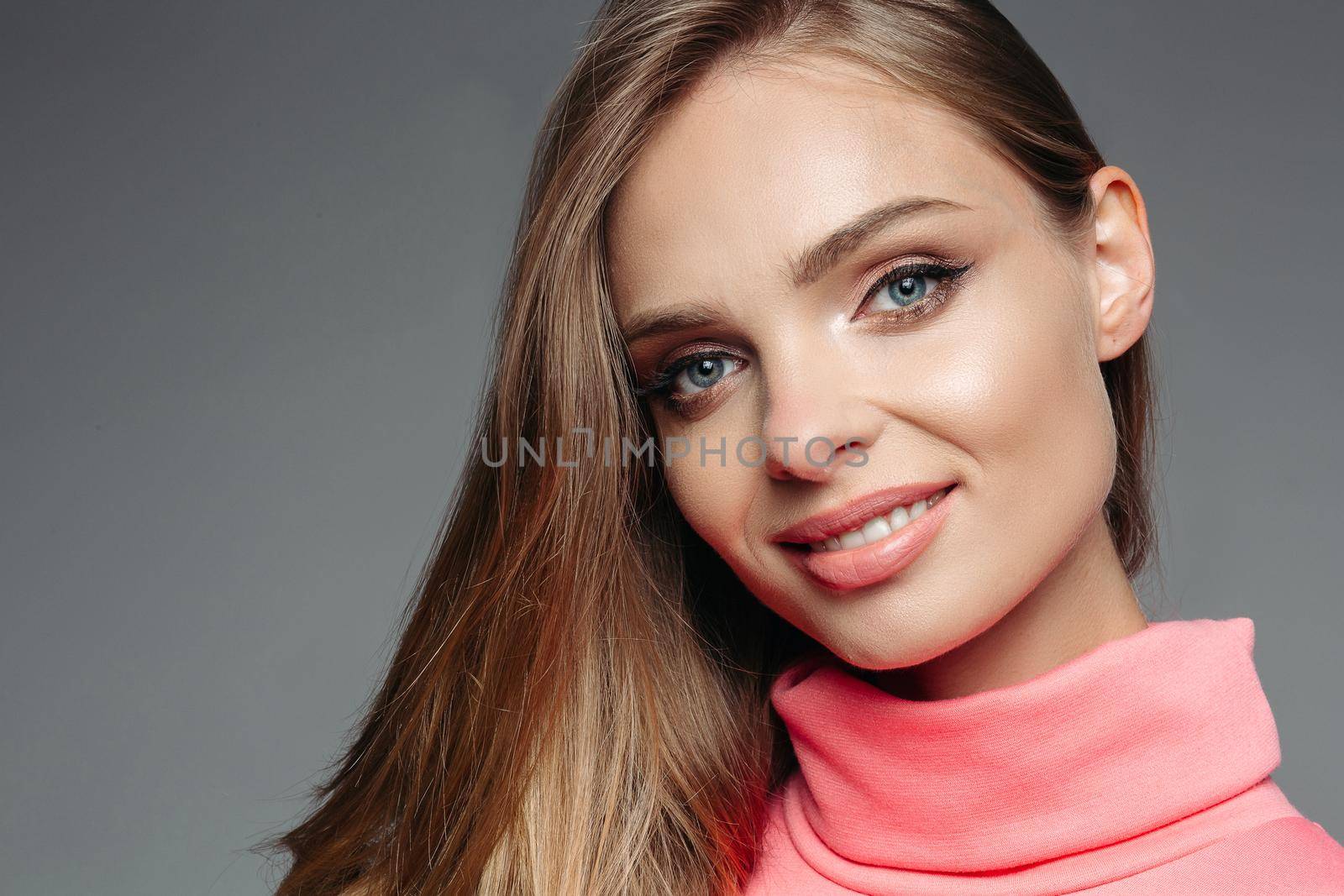 Portrait of pretty and gorgeous blonde woman in pink sweater with wavy volumed hair, sensuality smiling at camera. Girl with perfect make up after beauty salon. Gray studio background.