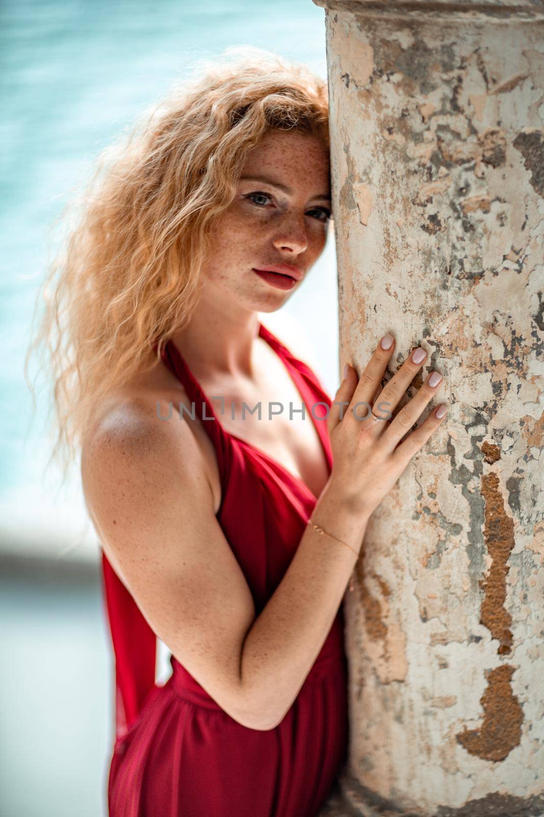 Outdoor portrait of a young beautiful natural redhead girl with freckles, long curly hair, in a red dress, posing against the background of the sea. by Matiunina