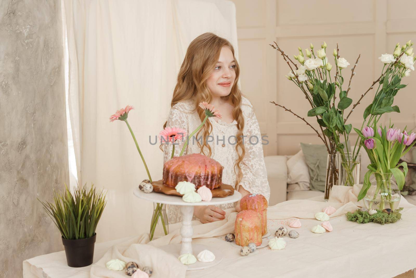A girl with long hair in a light dress is sitting at the Easter table with cakes, spring flowers and quail eggs. Happy Easter celebration