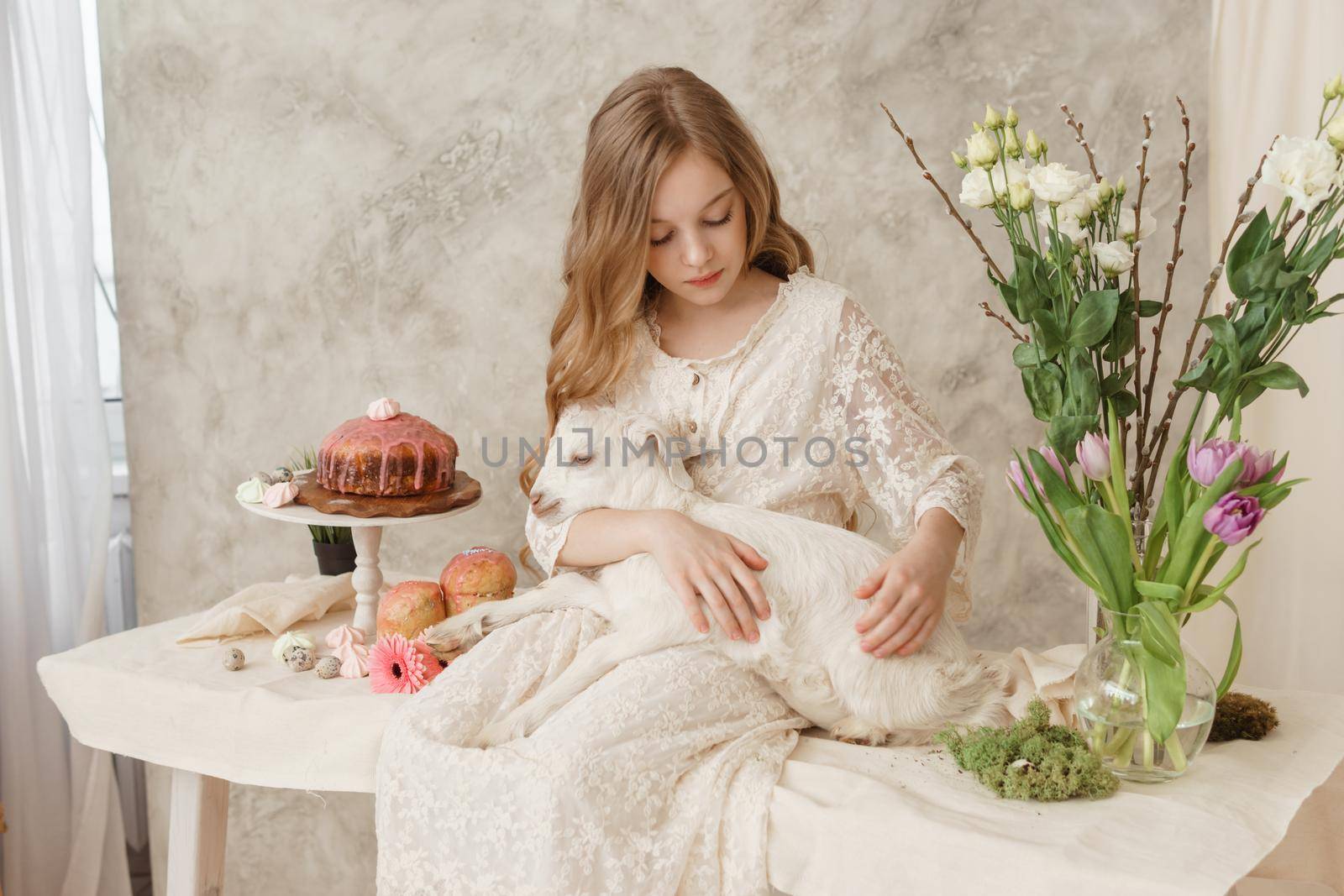 The girl is sitting at the Easter table with cakes, spring flowers and quail eggs and holding a white kid in her arms. Happy Easter celebration