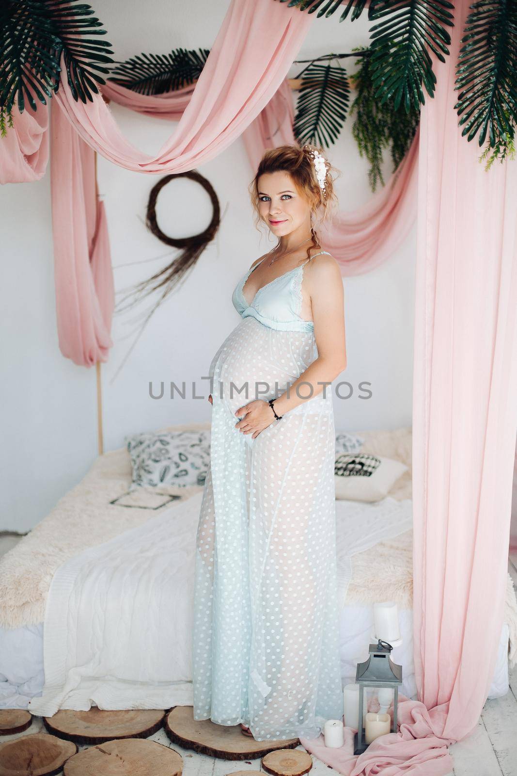 Above view for beautiful blonde pregnant woman, lying on bed, touching, holding hands on stomach, looking at camera. Pretty young mom wearing in blue expecting for her baby. Stylish home interior.