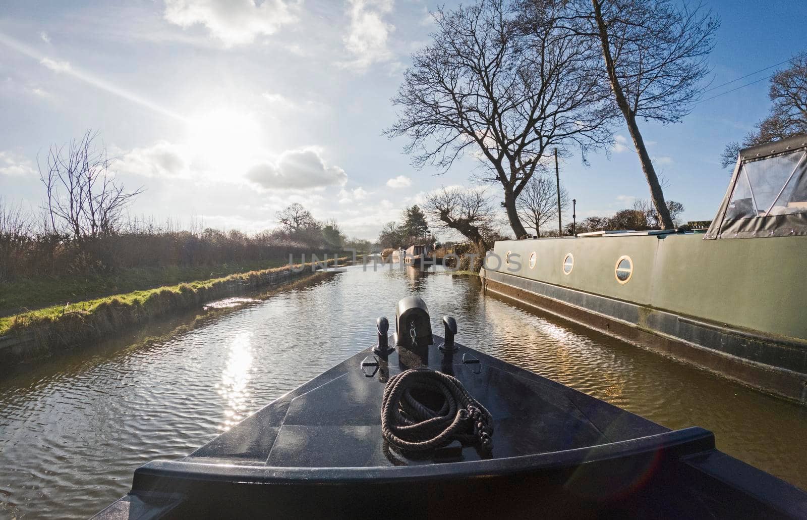 View from the bow of boat with narrowboats moored up in English rural countryside scenery on British tree lined waterway canal