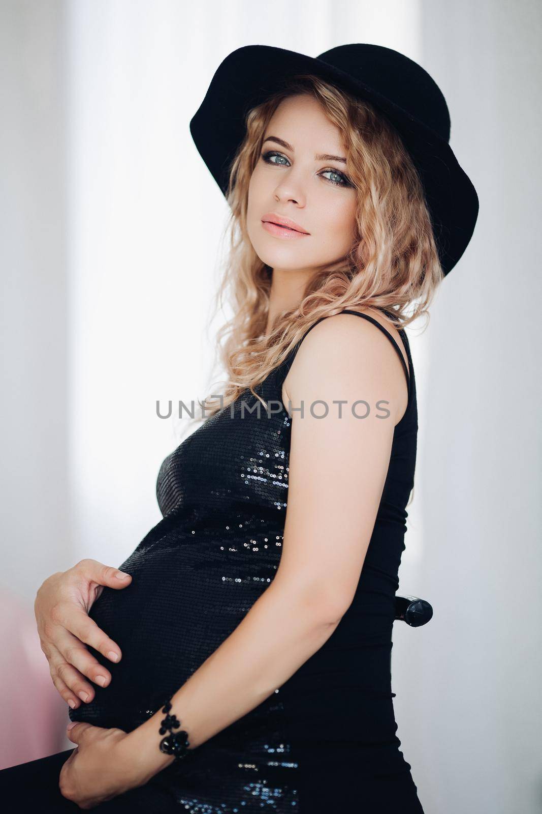 Studio portrait of attractive blonde woman with professional make up embracing her belly awaiting baby, wearing party black top and stylish black hat over white background.