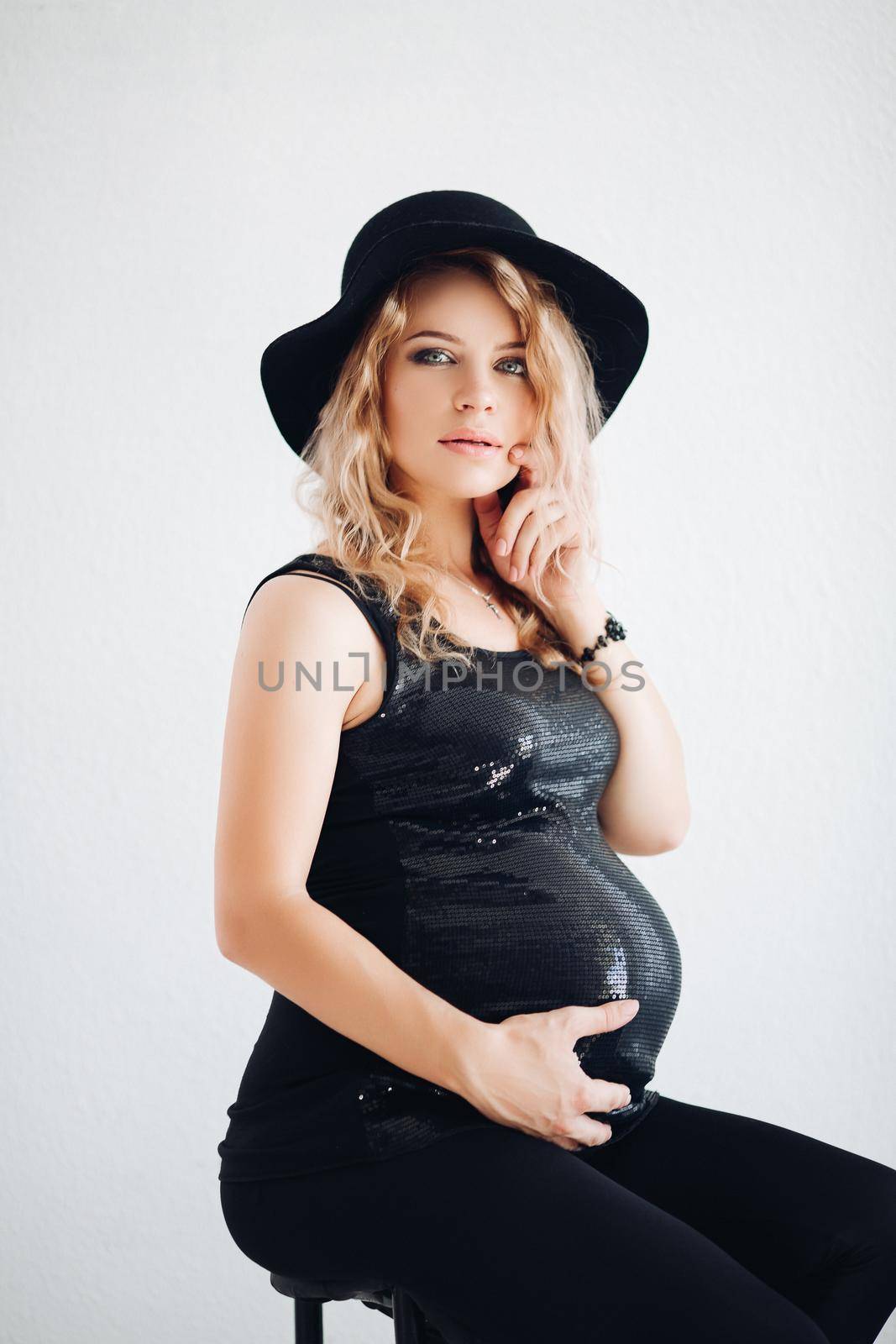 Studio portrait of attractive blonde woman with professional make up embracing her belly awaiting baby, wearing party black top and stylish black hat over white background.