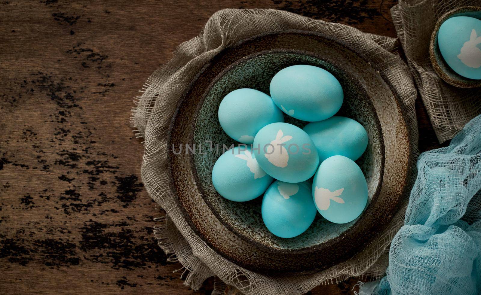 Unusual Easter on dark old background. Ceramic brown bowl with blue eggs with rabbit. Darkness, rays of sunlight shine on eggs. Concept of new life, rebirth. Rustic style. Top view, copy space